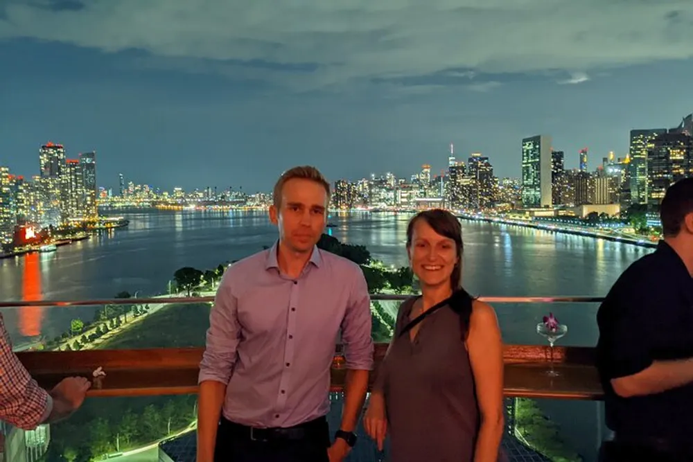A man and a woman are posing for a photo with a brightly lit city skyline at night in the background