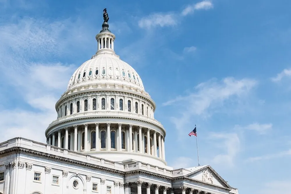 The image shows a close-up view of the United States Capitol dome with the American flag flying nearby against a partly cloudy sky