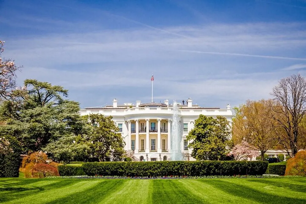 This image features the White House with its iconic facade green lawn and flowering trees under a clear blue sky