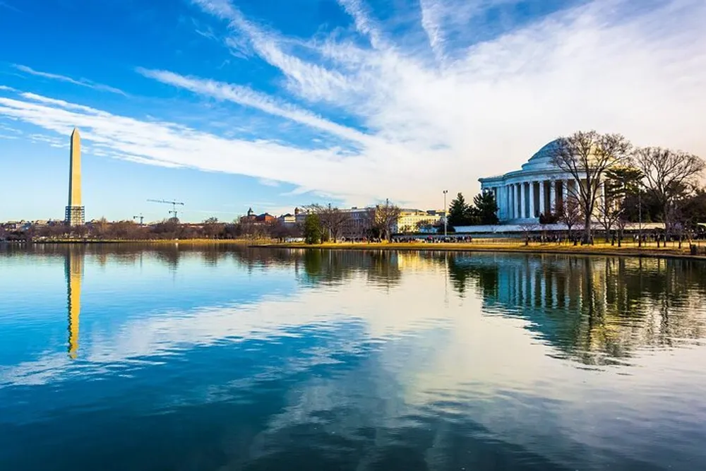 The image captures a serene view of the Washington Monument and the Thomas Jefferson Memorial reflected in the Tidal Basin under a blue sky with wispy clouds