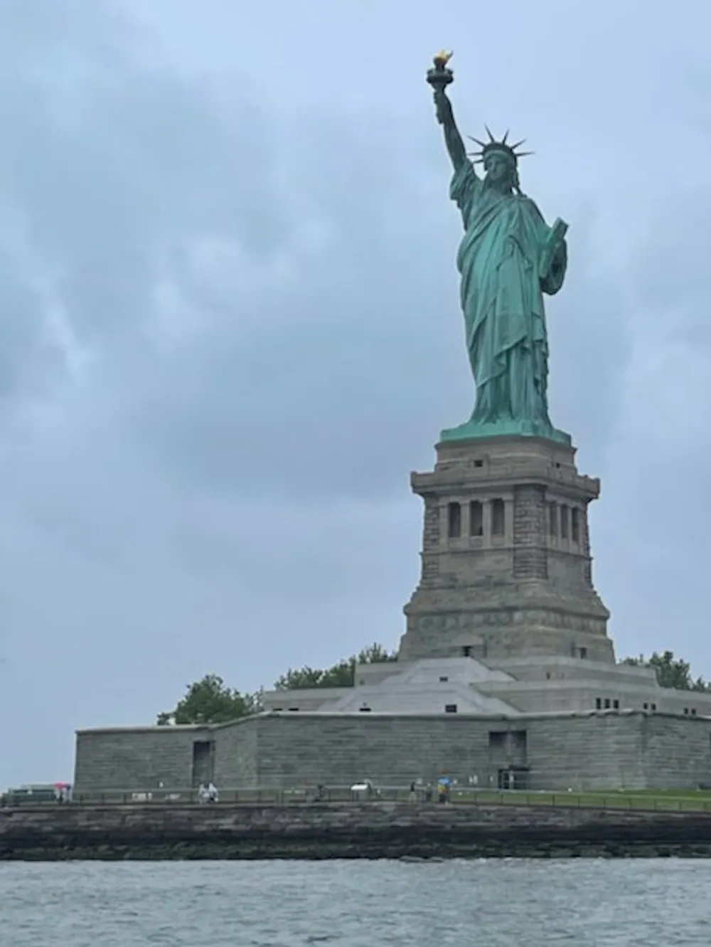 The image shows the Statue of Liberty on a cloudy day as viewed from the water with a focus on the statue and its pedestal