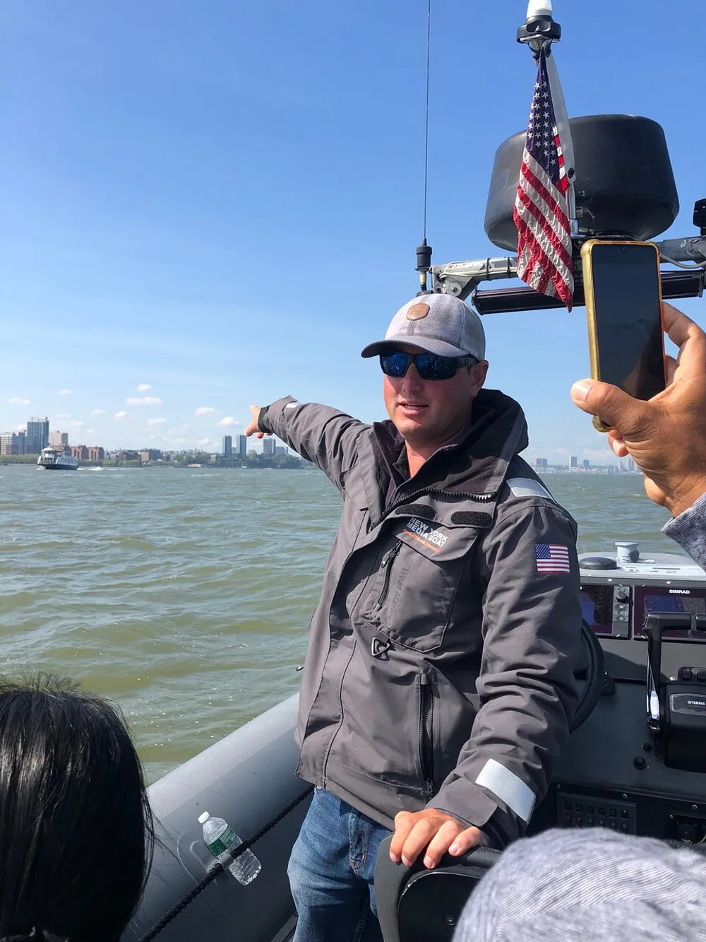 A person in sunglasses and a baseball cap is pointing towards something out of frame while on a boat as someones hand holds a smartphone possibly capturing a picture or video with an American flag prominently displayed in the background