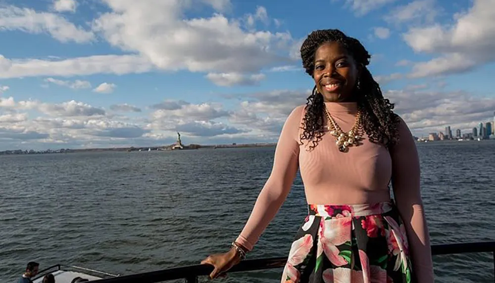 A smiling woman is standing on a boat with the Statue of Liberty and the New York City skyline in the background