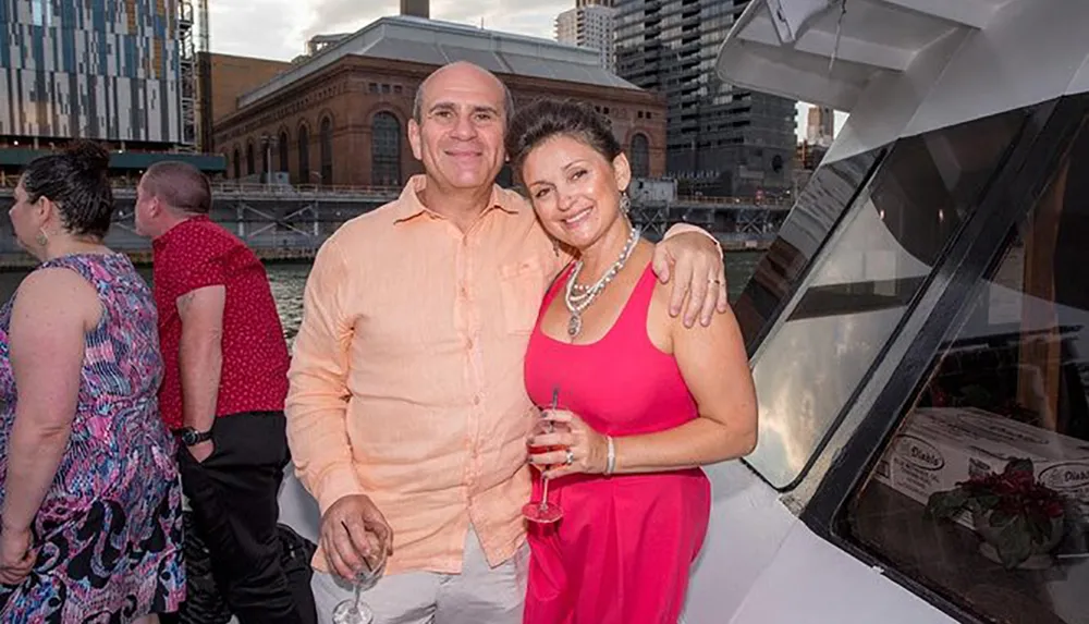 A man and woman are smiling for the camera while holding wine glasses on a boat with urban buildings in the background suggesting a social event or celebration