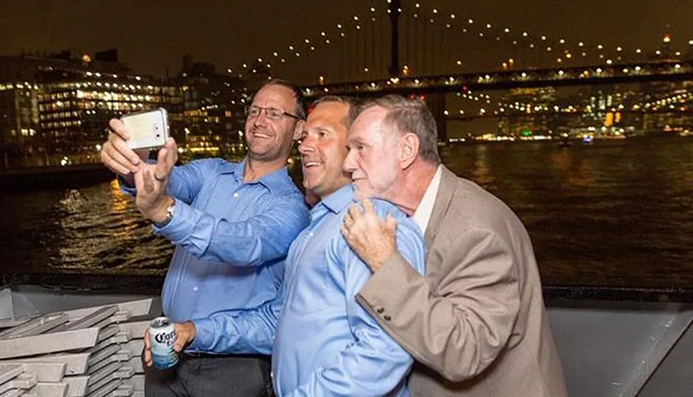 Three smiling men are taking a selfie together at night with a lit-up bridge and cityscape in the background